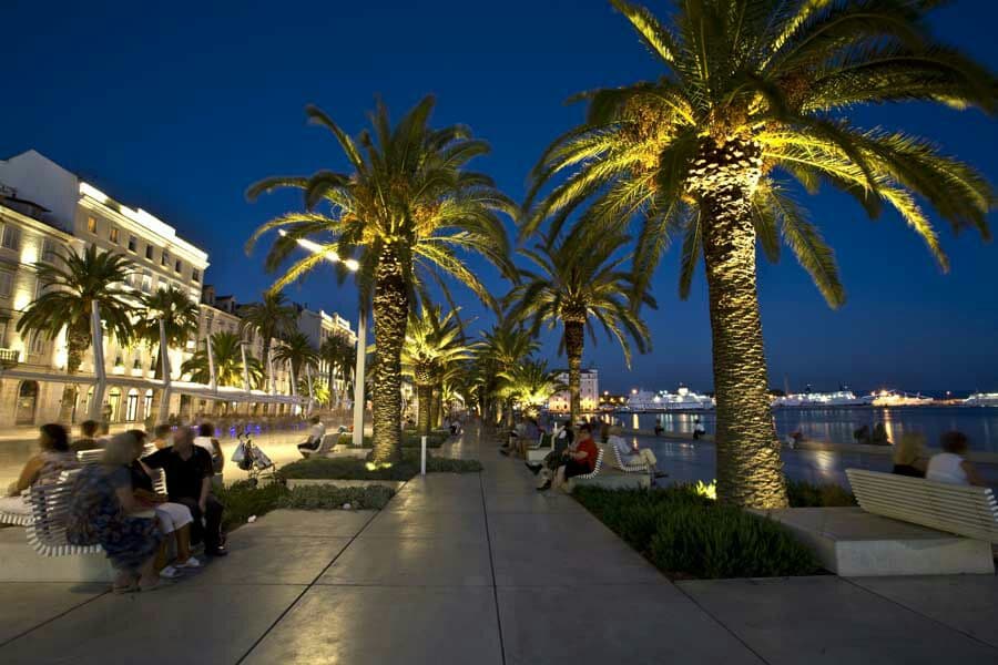 A paved walkway lined with illuminated palm trees creates a welcoming atmosphere at night. Benches are positioned along the path, where people can be seen enjoying the surroundings. The waterfront glimmers softly in the background, contributing to the scene's tranquil ambiance.