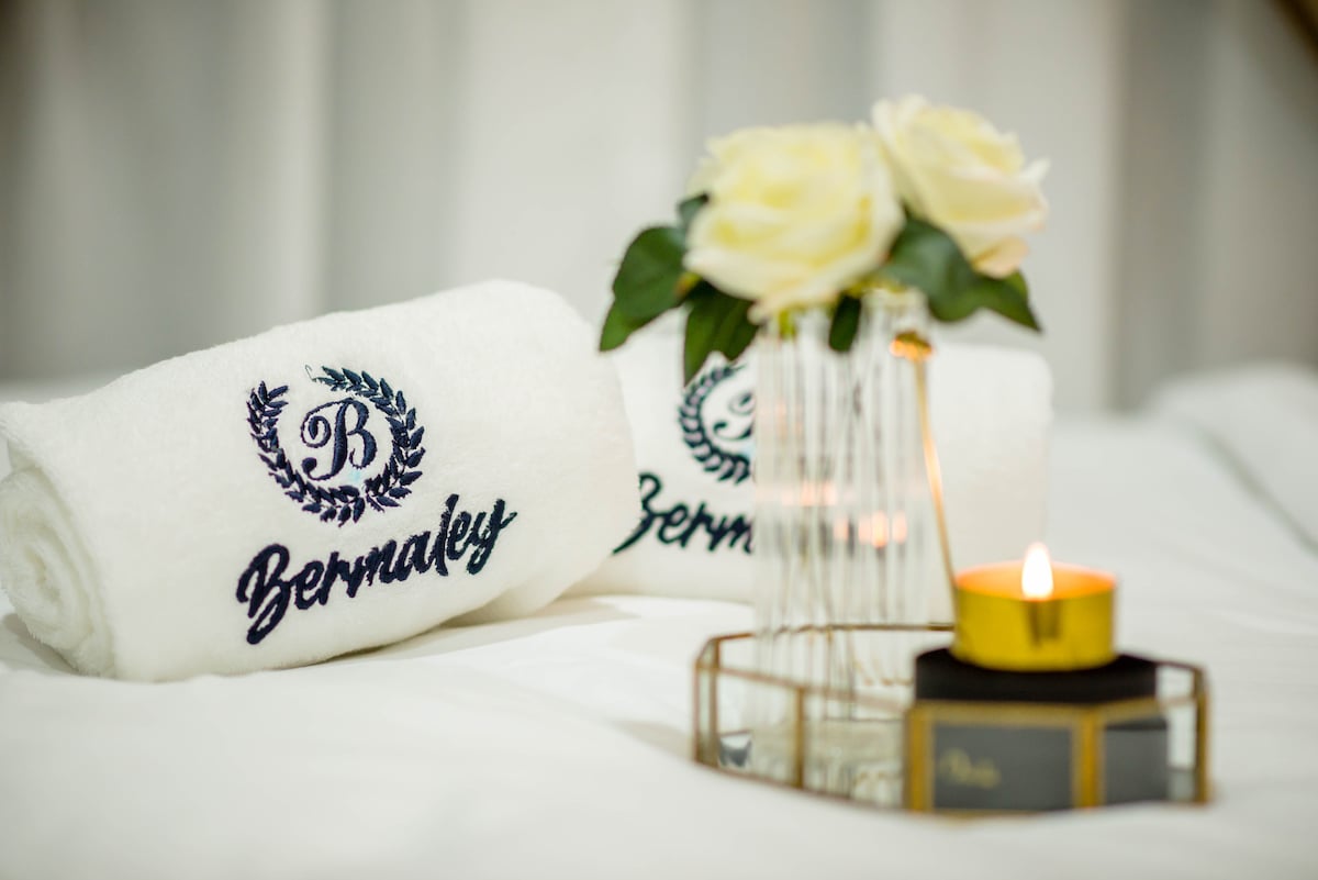 Two rolled towels with the Bermaley logo are displayed on soft white linens, accompanied by a small vase holding delicate white roses. A lit candle with a gold rim rests on a tray nearby, adding a touch of elegance to the setting.