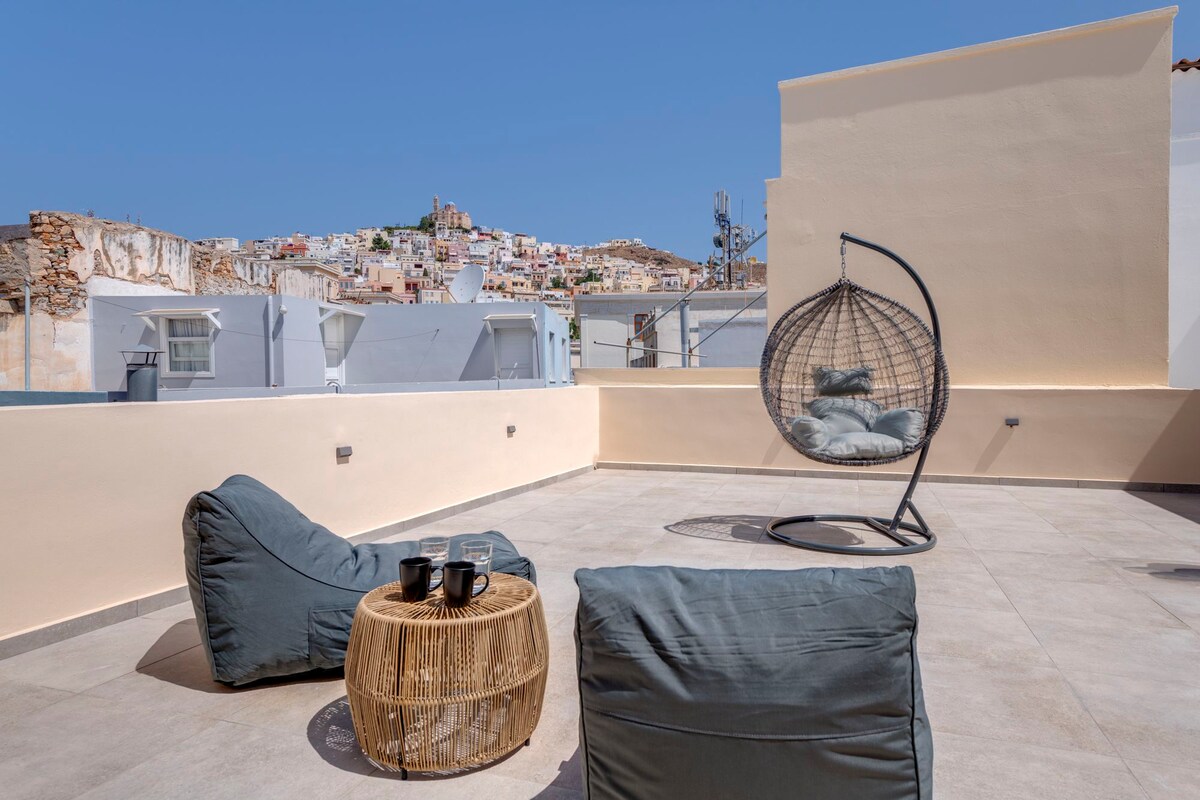 A spacious terrace features modern outdoor seating, including a hanging chair and bean bag chairs. A small wooden table holds two coffee cups. The backdrop showcases the scenic rooftops of Ermoupoli, with hills in the distance under a clear blue sky.