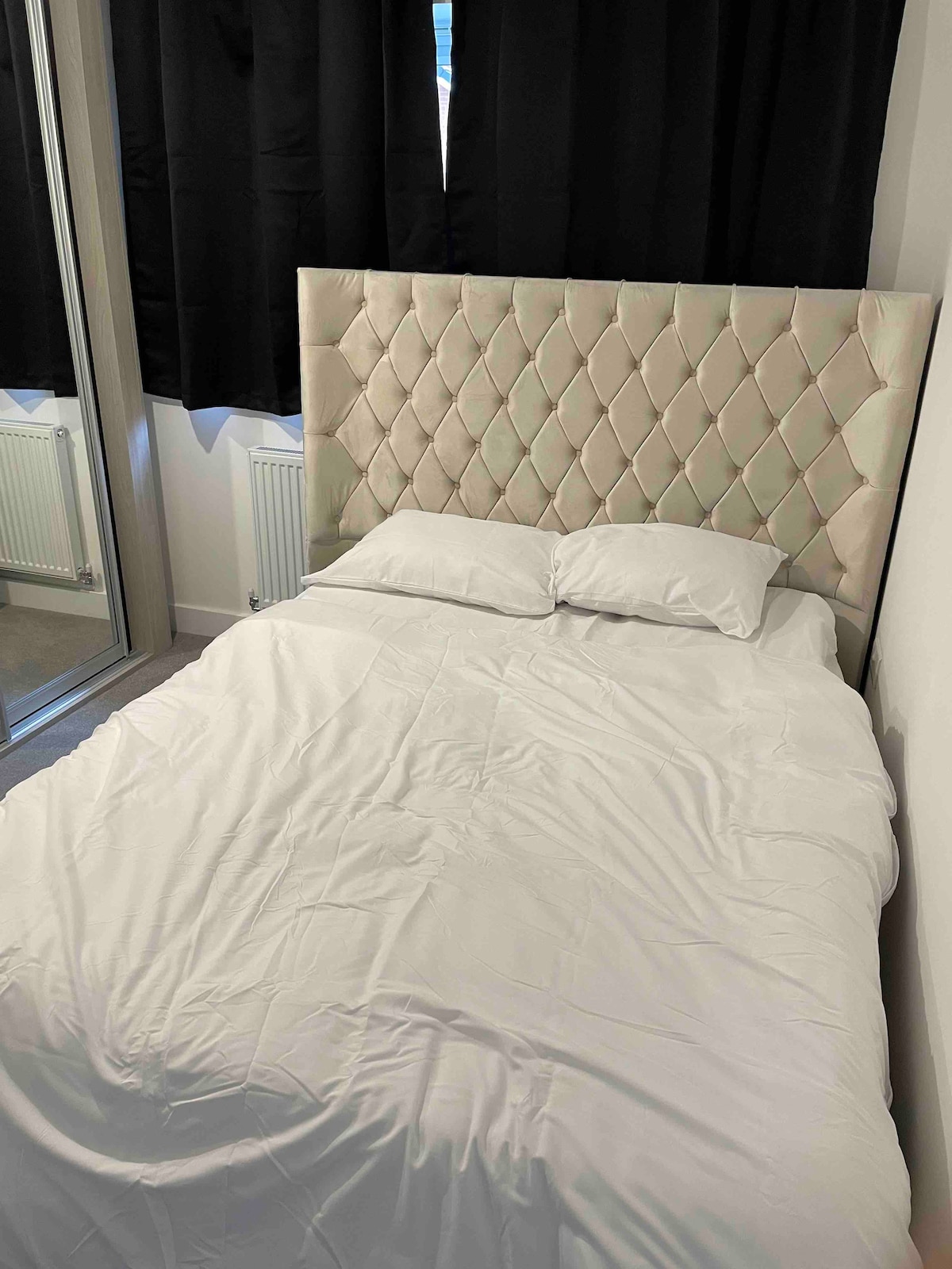 A cozy double bed is positioned against a tufted, light-colored headboard. Soft white linens cover the bed, and a pair of pillows rest on top. Dark curtains frame a nearby window, contributing to a serene atmosphere.