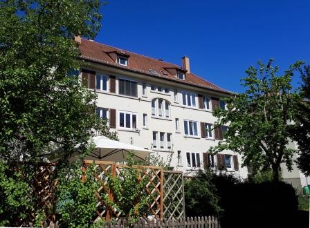 A three-story residential building is portrayed under a clear blue sky. The structure displays a mix of windows, with shutters framing each, and is surrounded by greenery, including trees and shrubs. A patio area can be seen featuring a canopy, enhancing the outdoor space.