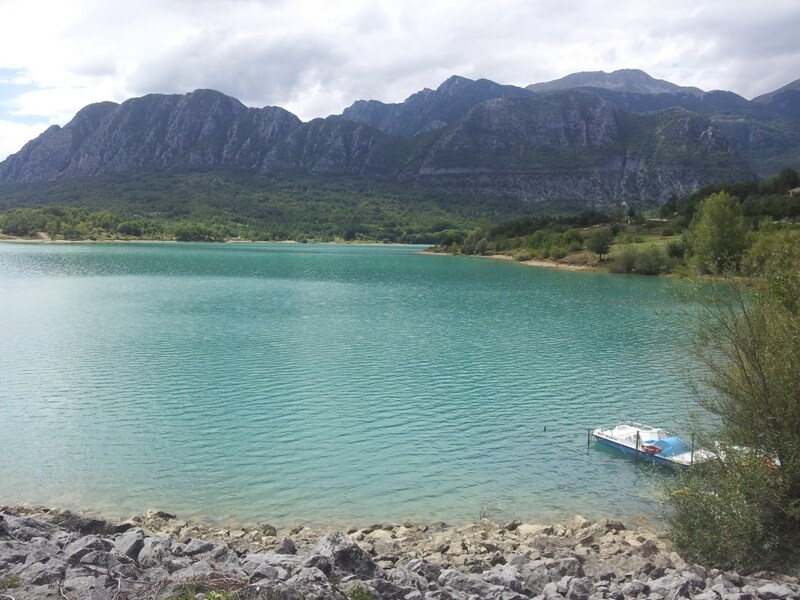 A serene lake is framed by rolling mountains under a partly cloudy sky. The water displays a vibrant turquoise hue, while a small boat is anchored along the rocky shore. Lush greenery lines the edges, enhancing the natural setting.