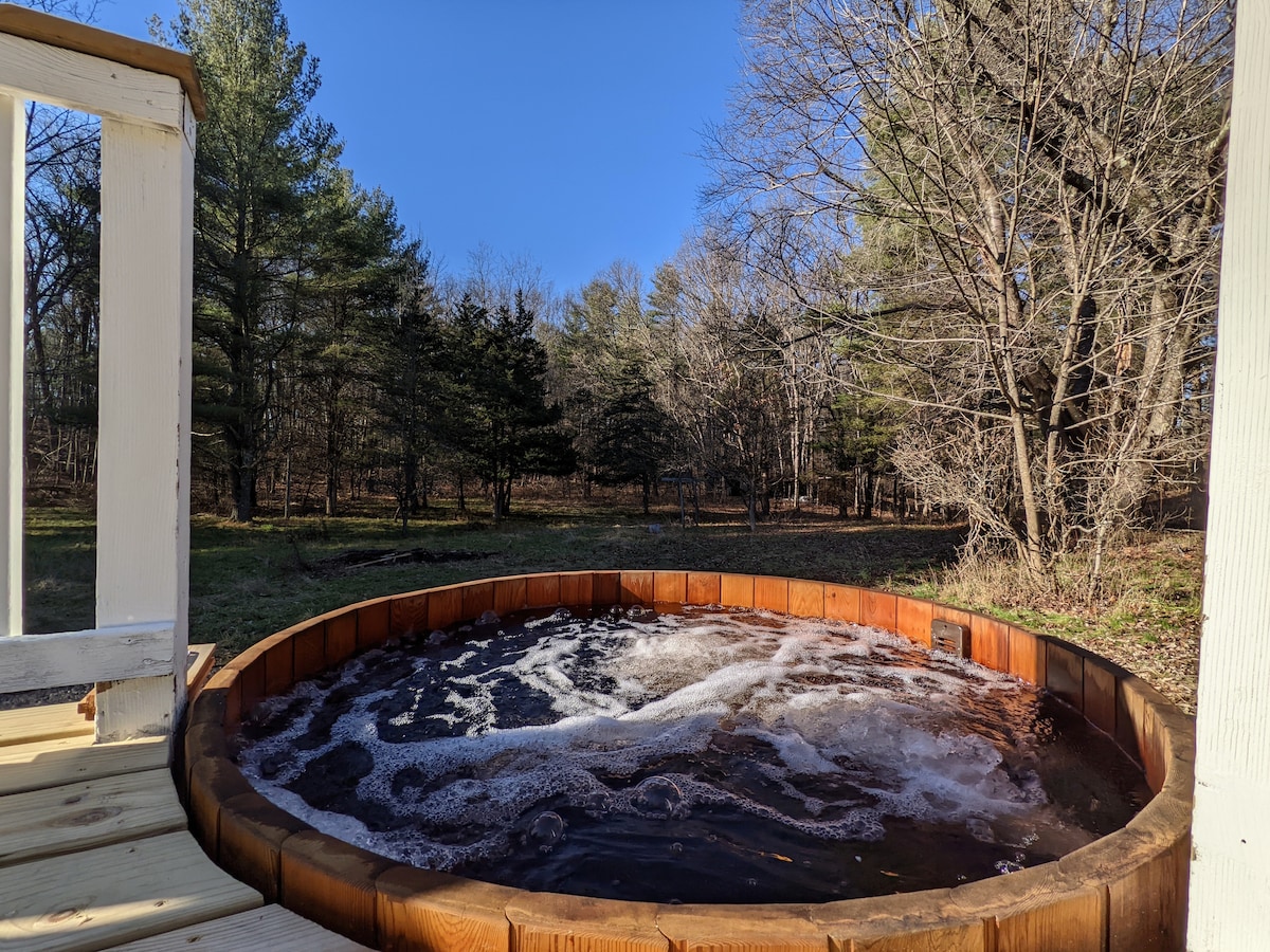 A spacious cedar hot tub sits peacefully outdoors, surrounded by a natural setting of trees and open space. Bubbles rise from the water, creating a relaxing atmosphere. Clear blue skies are visible above, enhancing the tranquil environment.