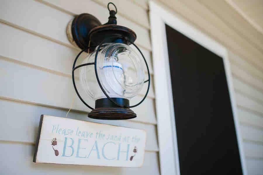 A decorative lantern is mounted on the wall beside a door, exuding a nautical theme. Below, a wooden sign displays the message, 'Please leave the sand at the BEACH,' emphasizing a coastal ambiance.