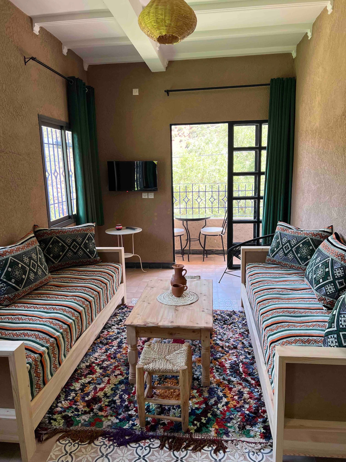 The living area features two patterned sofas with colorful cushions positioned around a wooden coffee table. A flat-screen TV is mounted on the wall, and a balcony offers outdoor seating with a view through large windows adorned with green curtains.