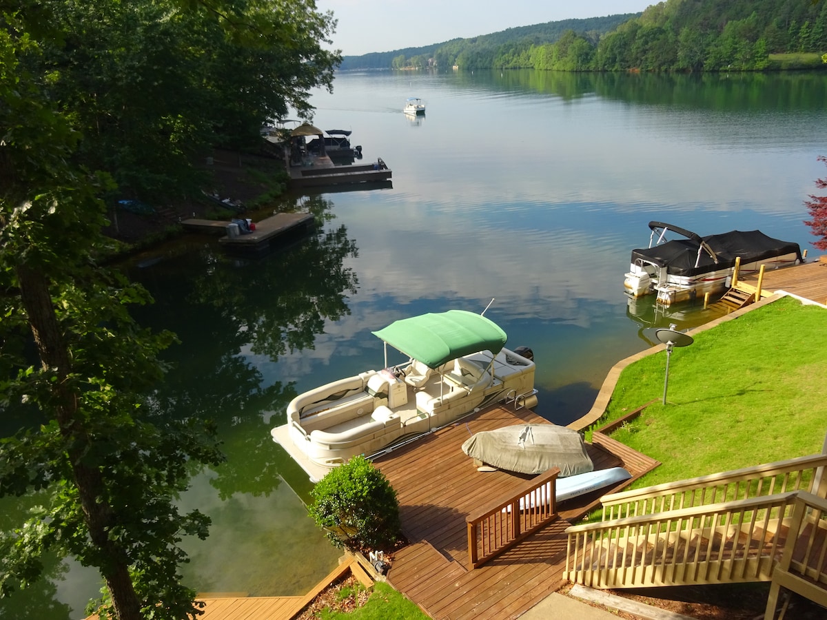 A serene lakeside view captures gently rippling water reflecting green trees. A pontoon boat with a green canopy is docked on a wooden platform. Surrounding areas feature well-maintained grass and additional boat docks, with a tranquil atmosphere enhanced by the calm surroundings.