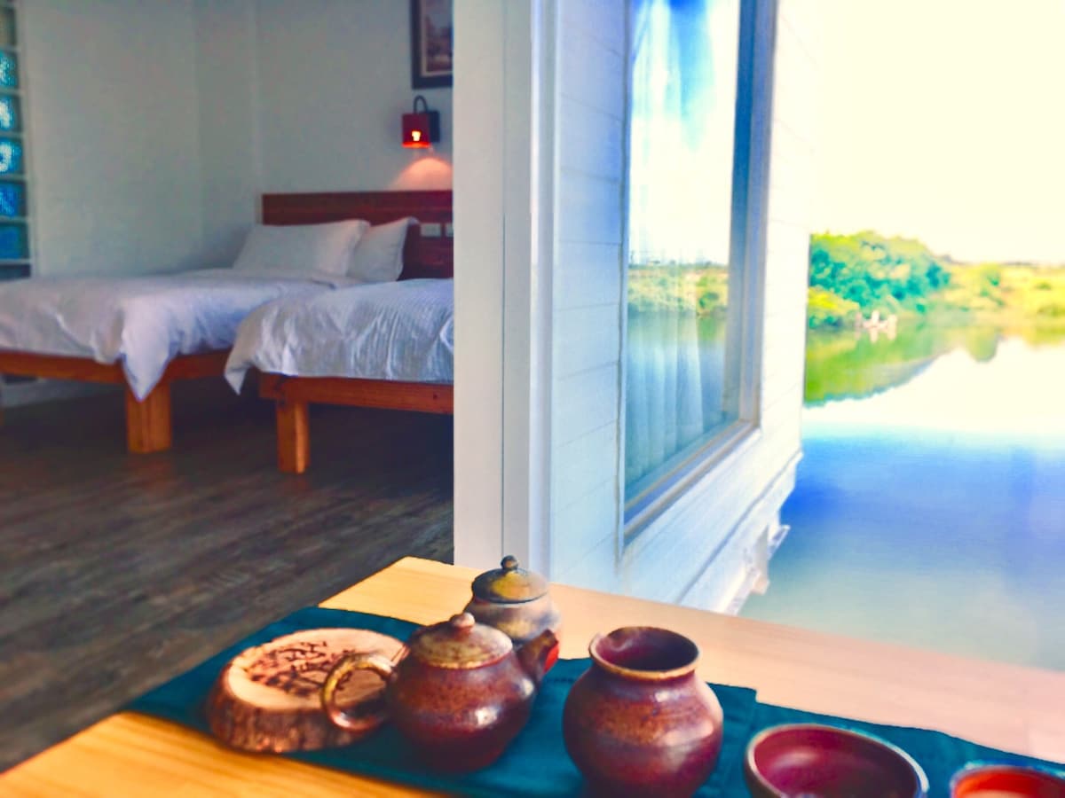 A serene space is featured with two spacious beds neatly made in white linens. In the foreground, a dining table displays traditional pottery and wooden trays, while a large window reveals a tranquil view of water and greenery outside.