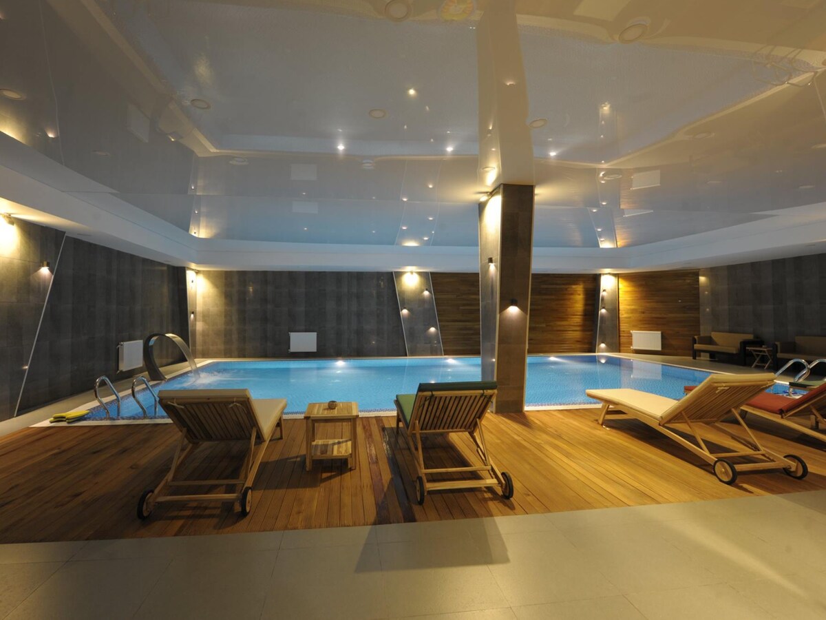 An indoor swimming pool area is highlighted by a reflective ceiling and subtle lighting. Lounge chairs are positioned around the pool's edge on wooden flooring. The space features a mix of textures, contributing to a relaxing environment.