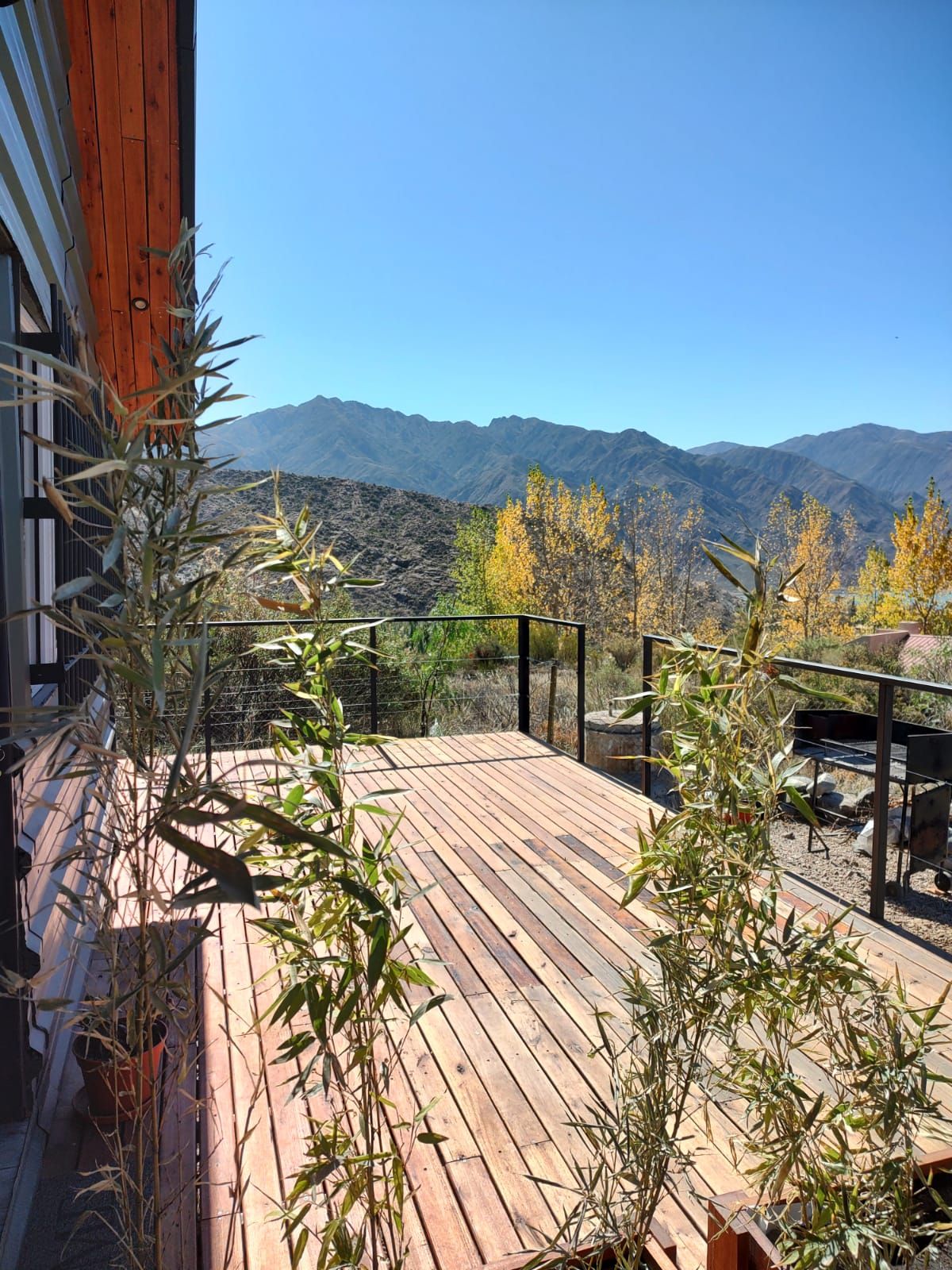 A spacious wooden deck overlooks a panoramic view of rolling mountains and clear blue skies, framed by tall bamboo plants on either side. The warm sunlight highlights the autumn foliage in the distance, creating a serene outdoor space for relaxation.