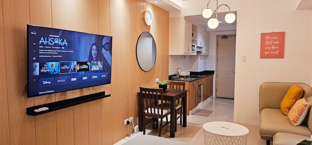 A modern living area is showcased, featuring a spacious sofa, a round coffee table, and a dining table with chairs. A large Smart TV is mounted on the wall, displaying streaming options. The kitchen, visible in the background, includes cabinets and essential appliances.