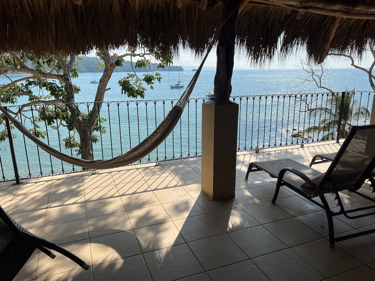 A balcony area is depicted, featuring a hammock suspended between two posts and lounge chairs positioned on tiled flooring. The expansive view of the shimmering ocean is framed by trees, and sunlight casts shadows across the space.