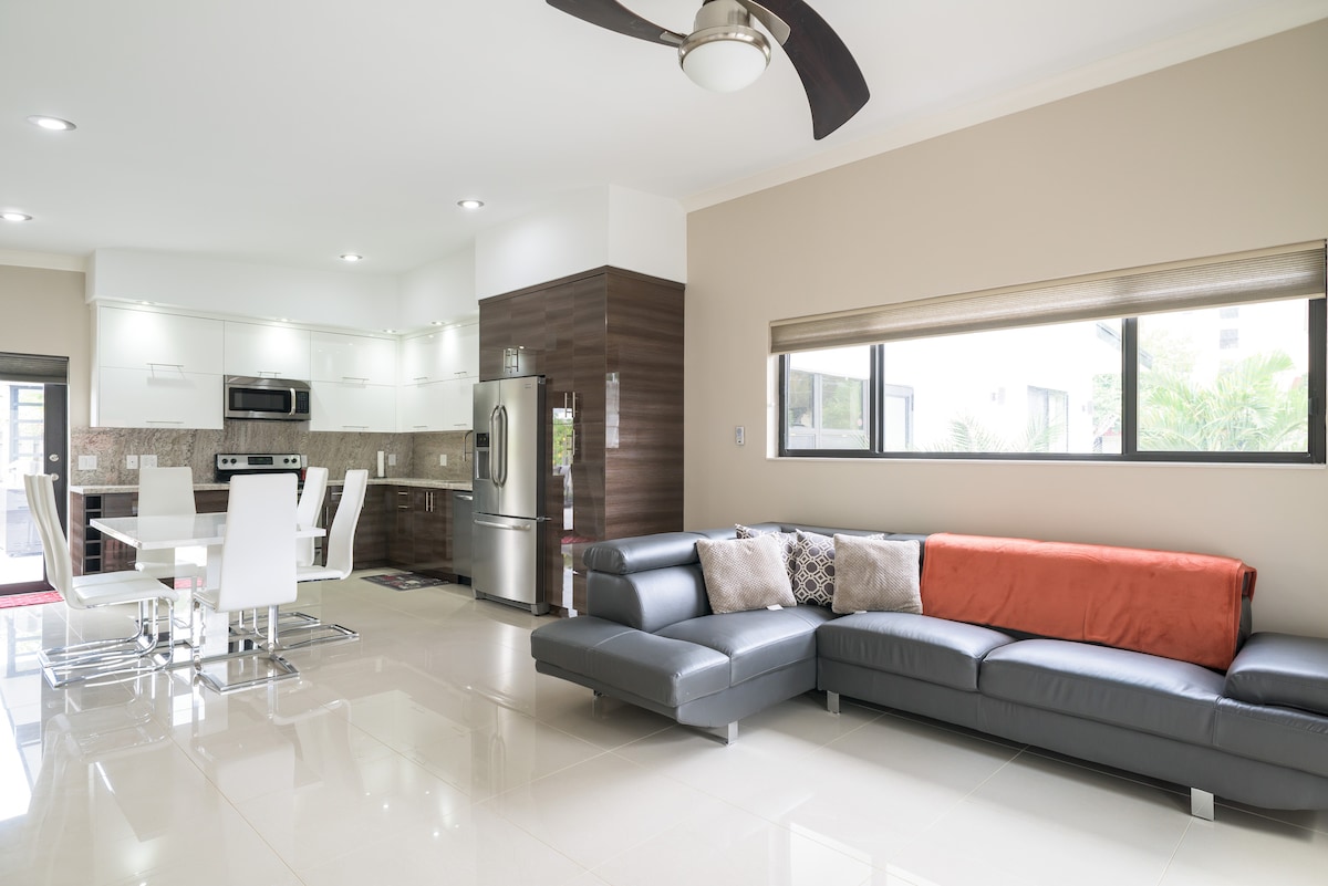 An open-concept living area features a modern kitchen with stainless steel appliances and a dining table for four. A sectional sofa in gray, complemented by an orange accent cushion, provides comfortable seating. Large windows allow natural light to fill the space, enhancing the neutral color palette.