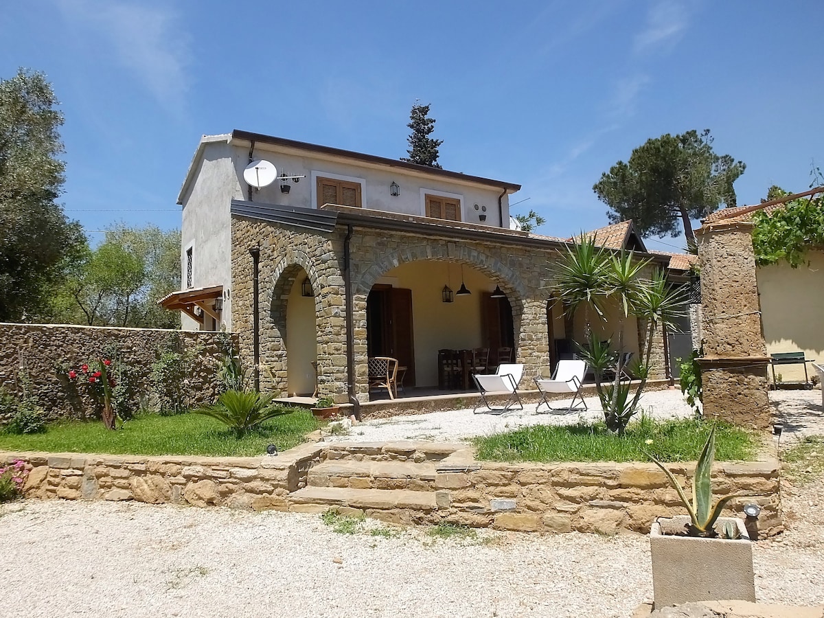 The exterior of the stone farmhouse is showcased, featuring a welcoming porch with arched columns. Two lounge chairs are positioned in the garden, surrounded by greenery. A pathway leading to the entrance is outlined by a stone wall, enhancing the inviting outdoor space.