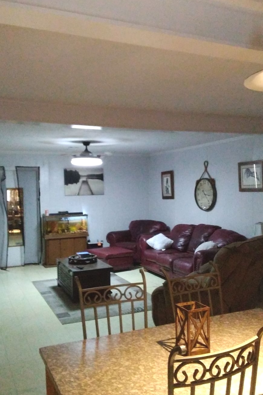 A spacious living area features comfortable leather seating arranged around a central coffee table. A wall clock and framed artworks are displayed, adding character to the room. A fish tank is visible in the corner, providing a bright focal point. Soft lighting enhances the welcoming ambiance.