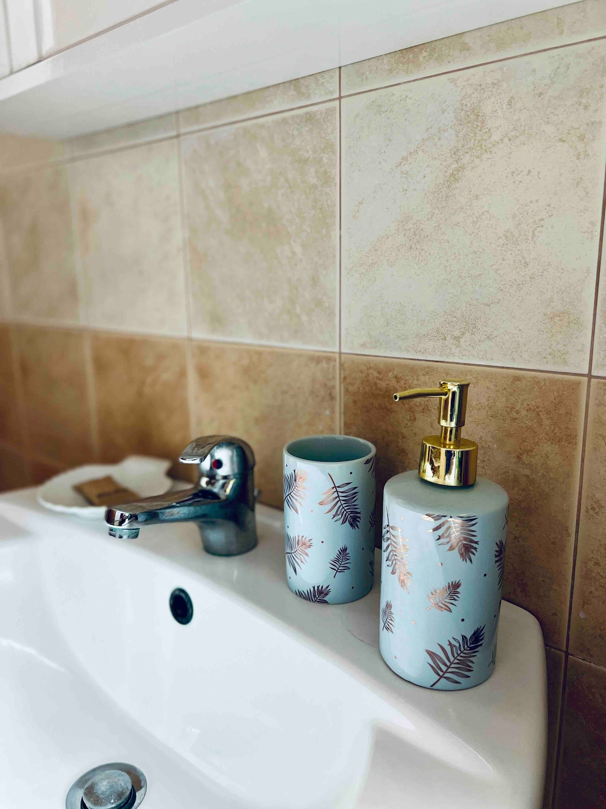 A bathroom sink area is displayed, featuring a chrome faucet and a soap dispenser with a gold pump. Two decorative containers in soft blue with leaf patterns complement the sink, with a small dish holding toiletries nearby.