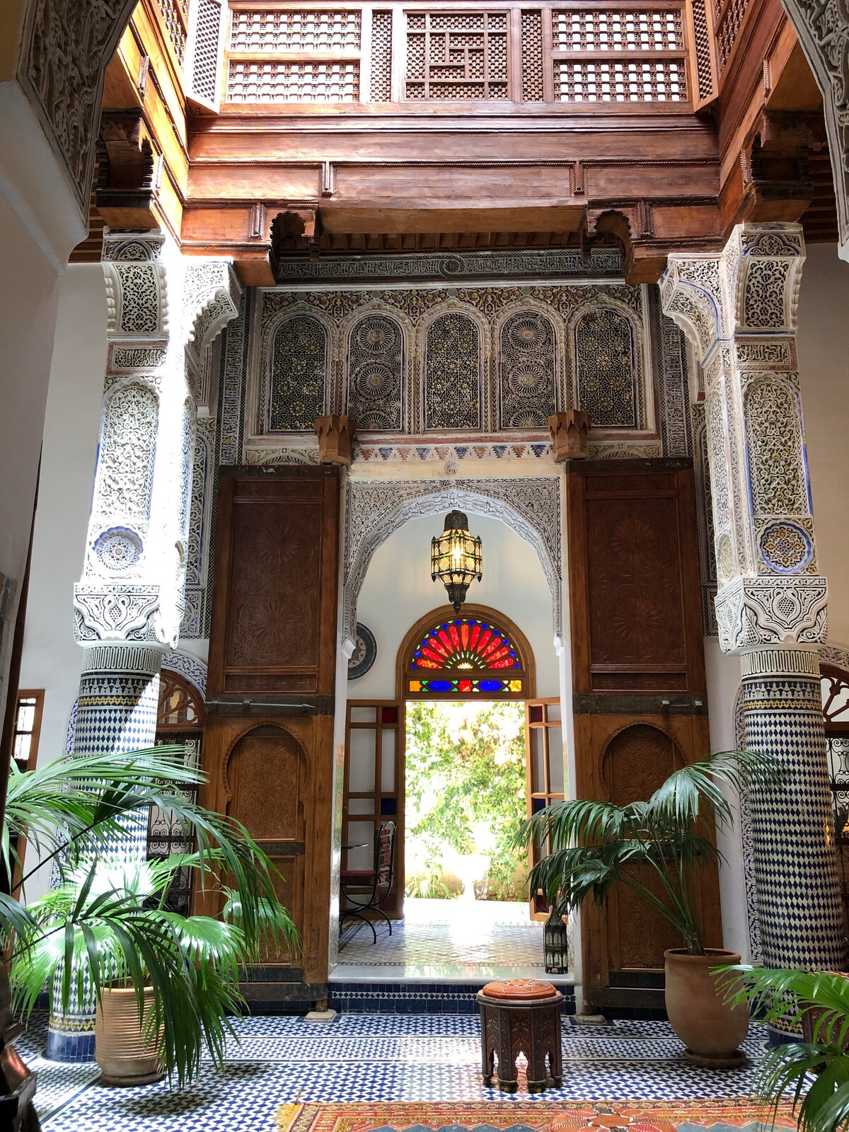 The entrance showcases ornate wooden doors with intricate carvings and a colorful stained glass window above, allowing natural light to filter in. Decorative tiles adorn the floor, complemented by lush greenery on either side, creating an inviting focal point that leads to the garden outside.