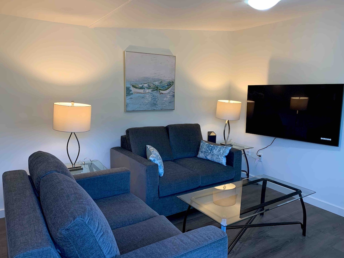 A comfortable living area is furnished with two gray sofas and a glass coffee table. Two lamps provide soft illumination, enhancing the space's ambiance. A large flat-screen television is mounted on the wall across from the seating, complemented by an art piece above the couch.