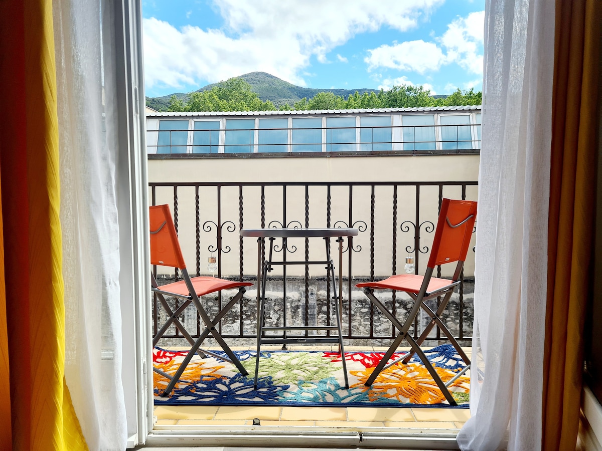 A balcony is visible through open French doors, featuring two orange folding chairs and a small table. The space is adorned with a colorful rug, and a view of the mountains is framed by the lush greenery of the surrounding area.