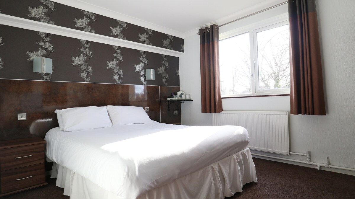 A bright bedroom is presented with a spacious bed dressed in white linens. Dark floral wallpaper complements the wooden headboard. A bedside table sits beside the bed, and a window allows natural light to fill the room, enhancing its welcoming ambiance.