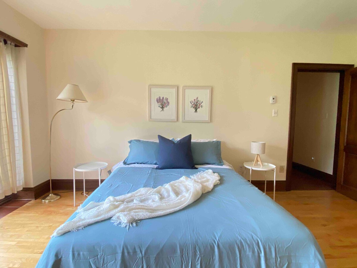 A well-lit bedroom features a spacious bed adorned with a soft blue cover. Two side tables flank the bed, one holding a lamp. Light filters through sheer curtains, while floral prints decorate the wall. A cozy blanket is casually draped over the bed.