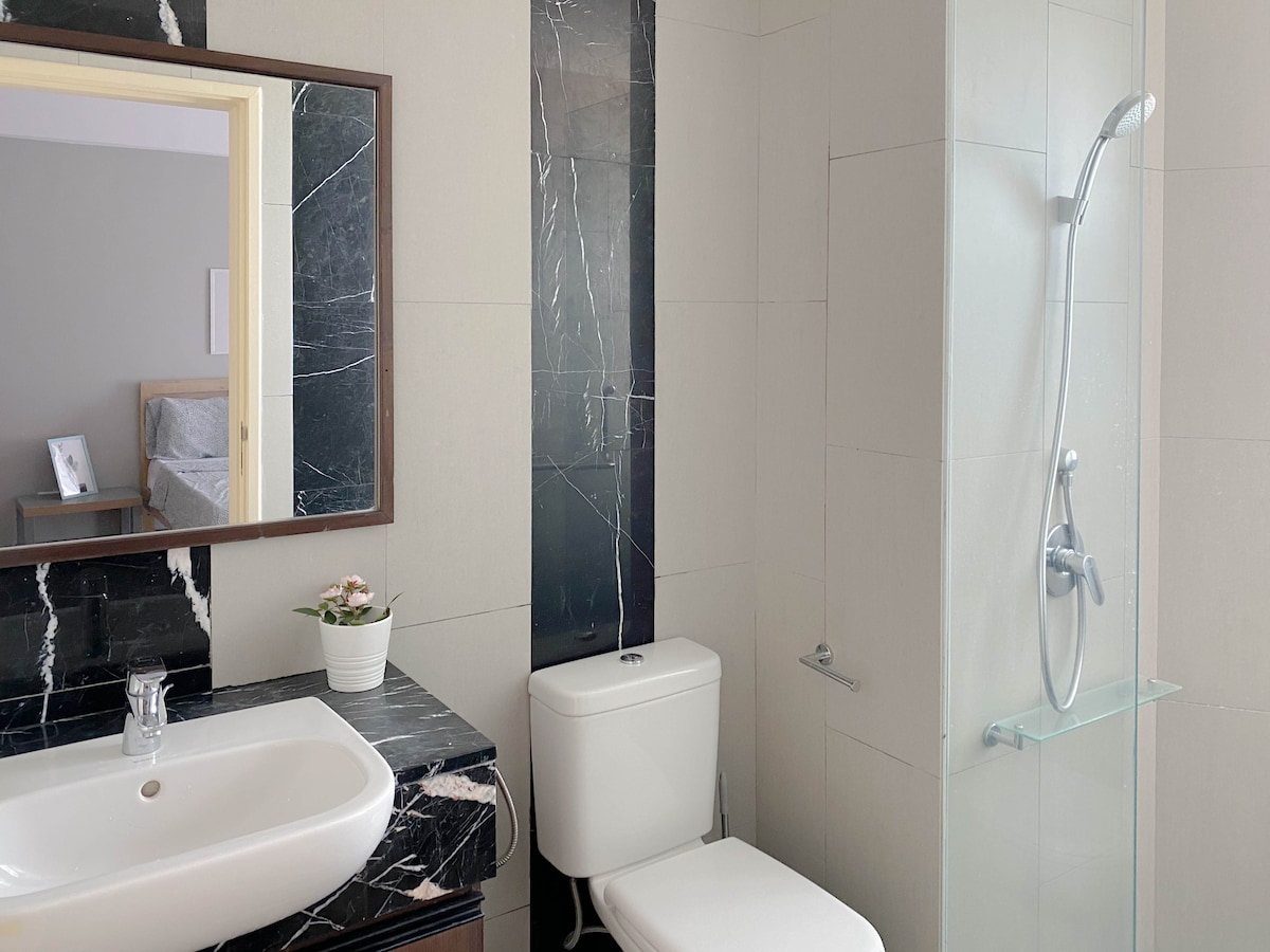 The bathroom features modern fixtures, including a sleek shower with glass partition and a wall-mounted faucet. A contemporary vanity with a dark marble countertop is accompanied by a round mirror. A potted plant adds a touch of greenery to the space.