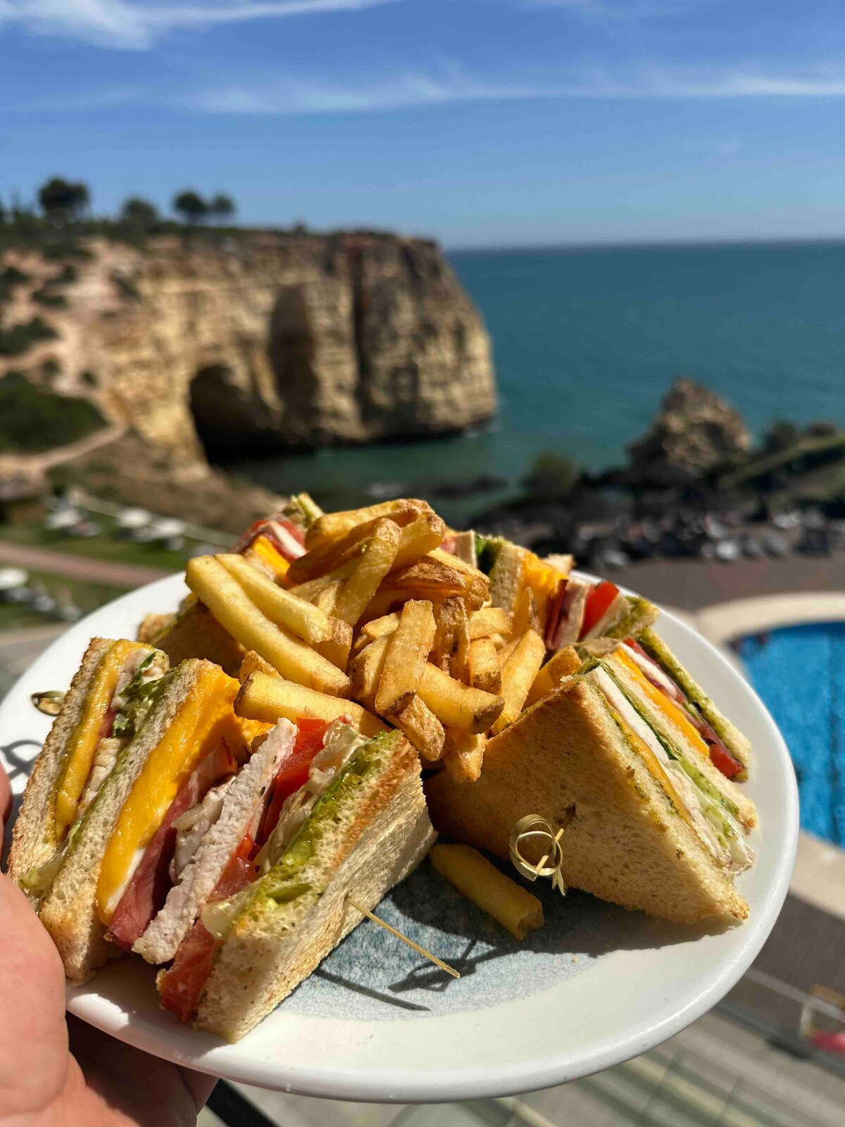 A plate is displayed featuring a layered sandwich with various ingredients, accompanied by a serving of golden french fries. The background reveals a stunning coastal view, with cliffs and azure water under a clear blue sky.