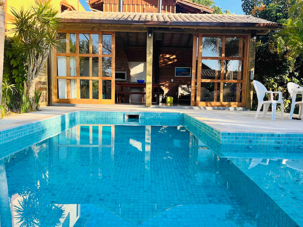 A tranquil saltwater pool reflects sunlight, surrounded by a wooden deck. The house features large glass doors that open to a spacious outdoor area, where tropical plants create a relaxing atmosphere. White chairs are positioned nearby, inviting guests to enjoy the serene environment.