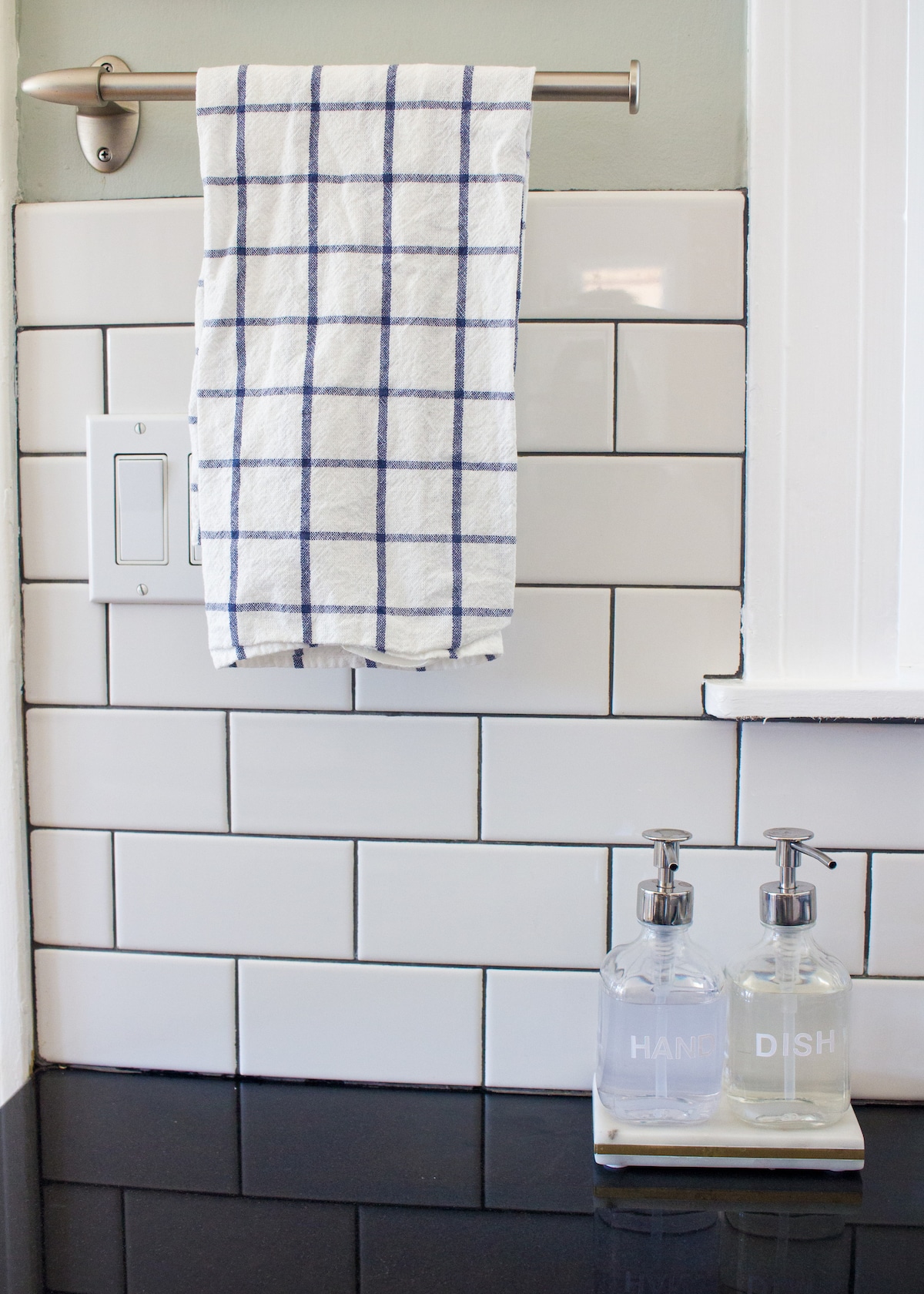 A clean kitchen area features a hanging dish towel with a classic checkered pattern on a silver rod. Below, two glass soap dispensers labeled 'HAND' and 'DISH' are positioned on a tray against a sleek black countertop, complemented by white subway tile backsplashes.