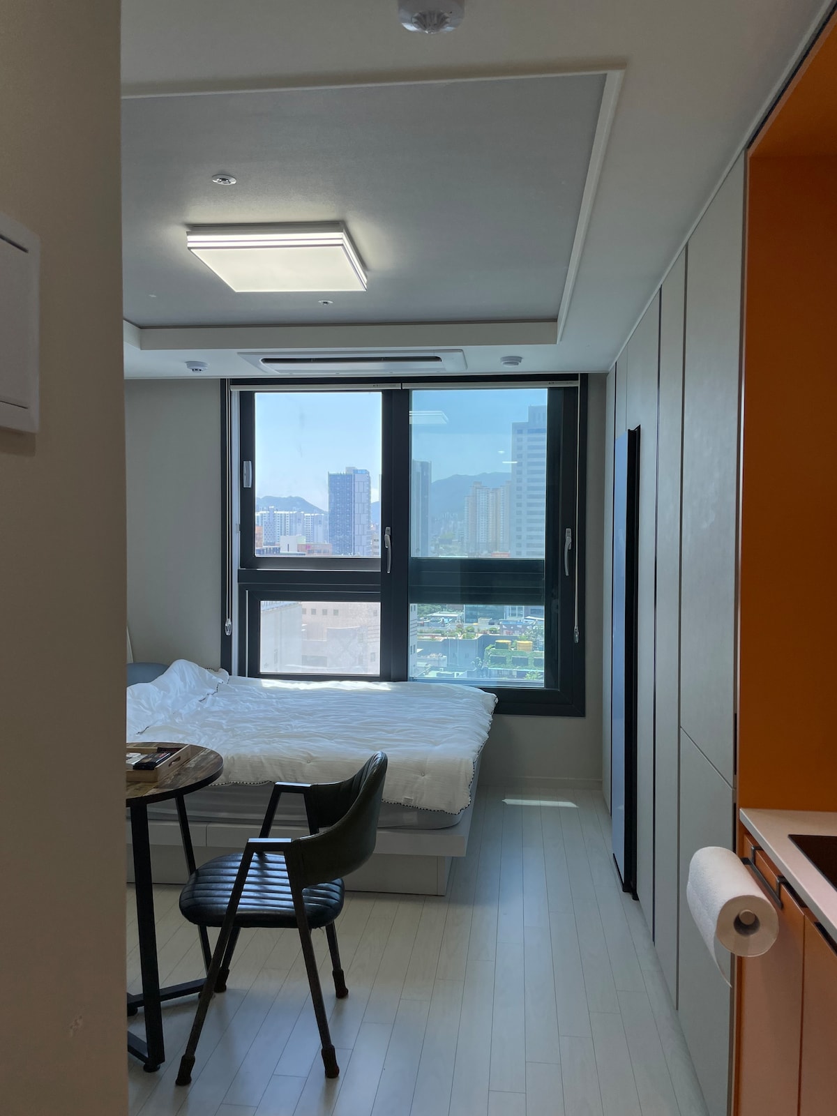 A well-lit room features a spacious bed with white bedding and a window that offers a city view. A small table and a black chair are positioned nearby, while an orange accent wall adds a touch of color to the otherwise neutral decor.