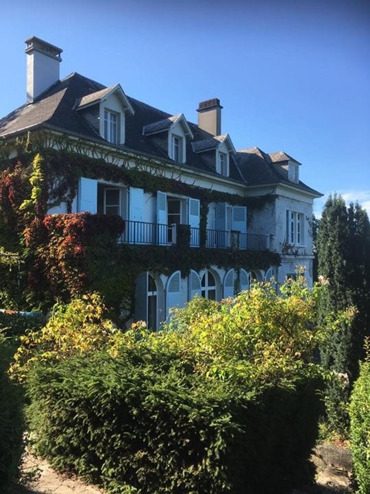 The elegant Maison de Maitre features a classic façade adorned with climbing greenery. Shuttered windows and a covered balcony add charm, while the surrounding garden includes lush shrubs and vibrant plants. The structure is set against a clear blue sky, emphasizing its grandeur.