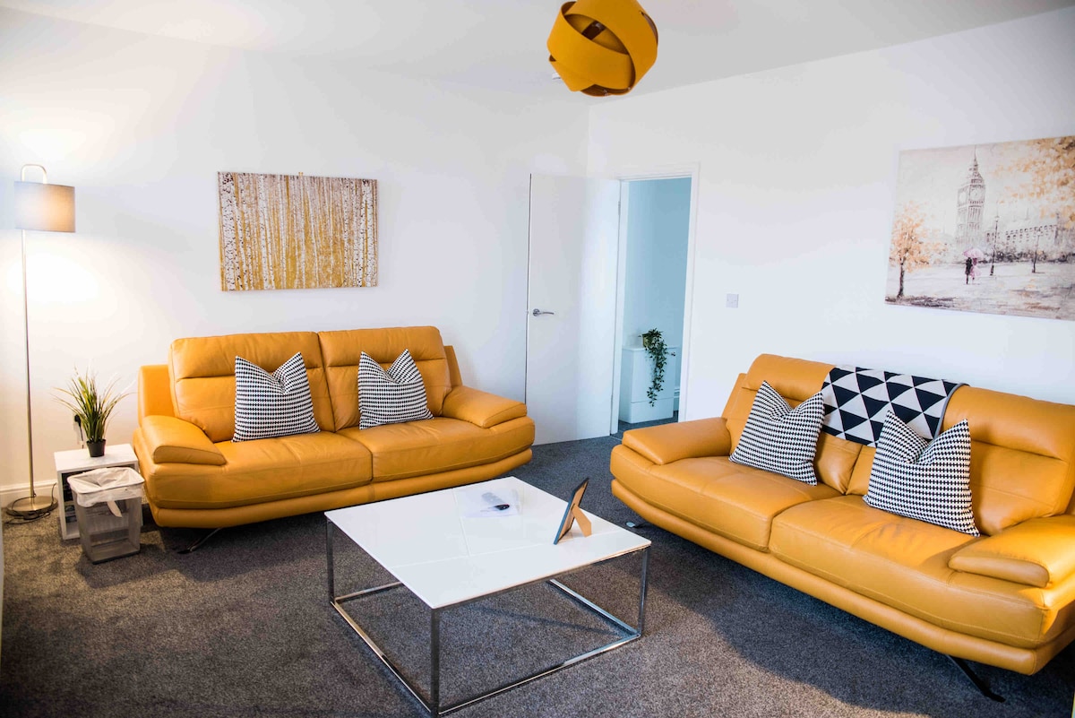 A bright living area features two yellow sofas with patterned cushions, set around a white coffee table. A contemporary lamp stands beside one sofa, while a piece of wall art depicts a tree-lined street. A doorway leads to another room in the background.