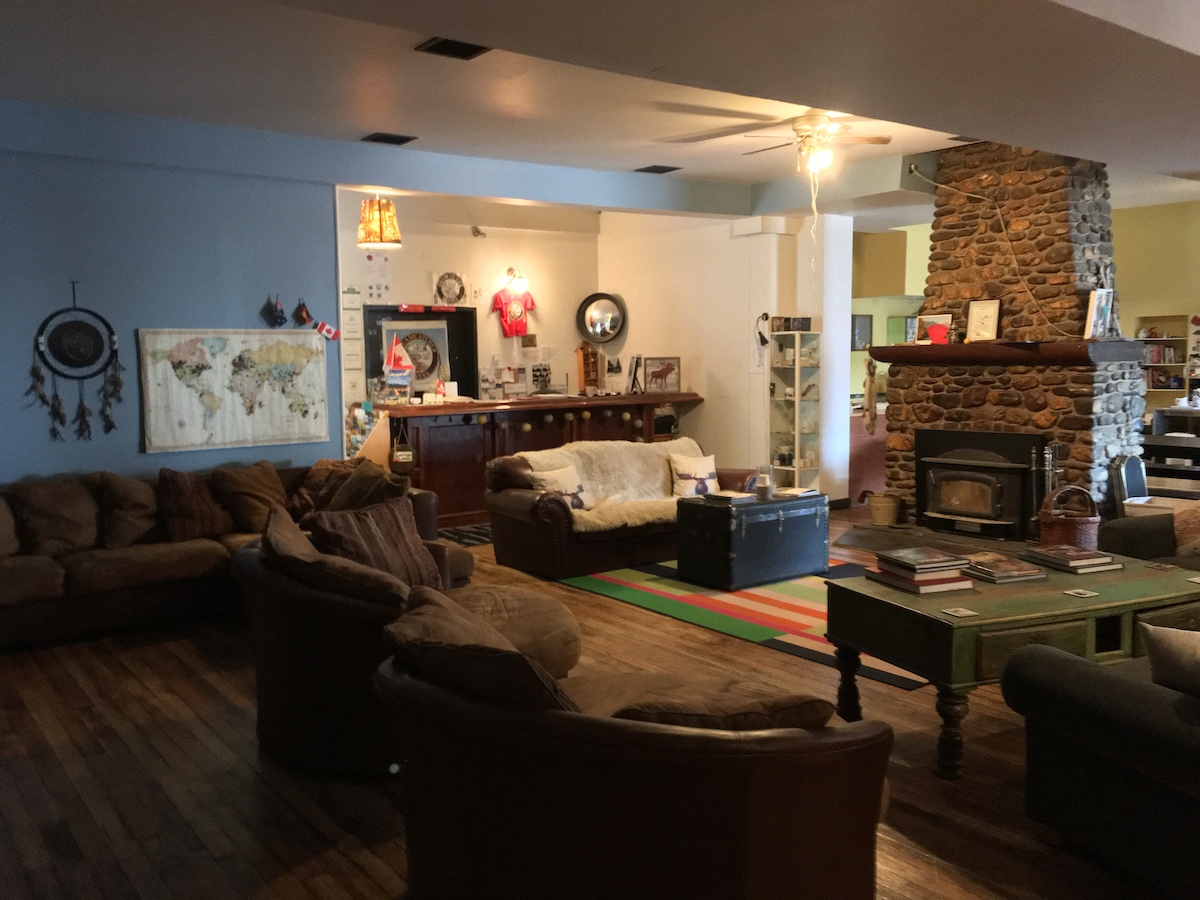 A spacious common area features comfortable seating arranged around a central stone fireplace. A warm rug is layered over hardwood floors, while a colorful table holds various items. Wall decor includes a world map and unique artwork, creating an inviting space for socializing.