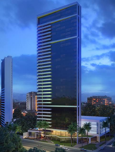 The tall building features a modern facade with reflective glass, capturing the evening sky. Palm trees are positioned in front, complementing the sleek design. The illuminated lobby area suggests a welcoming entrance, while the surrounding cityscape is visible in the background.