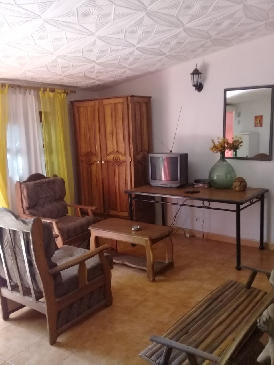 A cozy living area is presented, featuring a wooden wardrobe and a table with a vase of flowers. Two comfortable chairs are positioned around the table, and an older television stands against the wall. Natural light filters through sheer curtains, enhancing the space.
