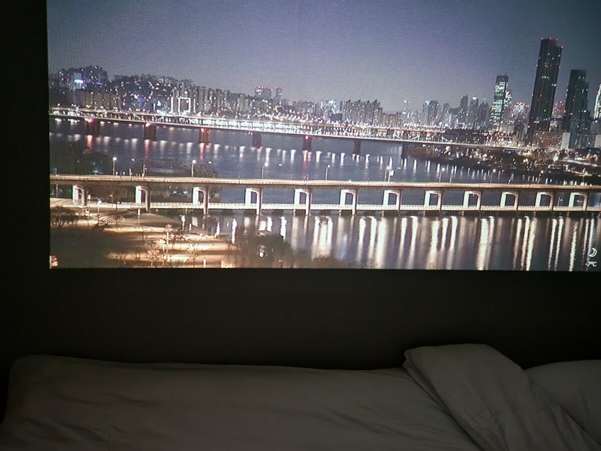A large image is projected onto the wall, displaying a serene night view of a city skyline. The water reflects the lights of the buildings, while a bridge spans across the foreground. A comfortable, unmade bed is positioned below the projection.