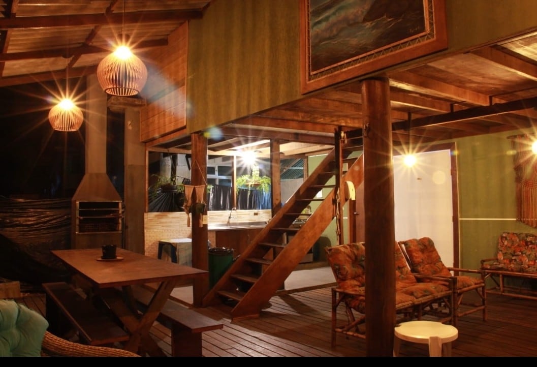 The spacious outdoor deck is furnished with a wooden dining table and colorful seating arrangements, offering a relaxed atmosphere. A staircase leads to the upper level, while a grill is positioned nearby. Soft lighting accentuates the warm tones of the wood and decor.