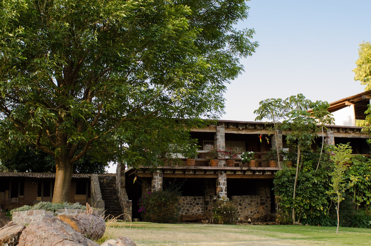 A rustic two-story building is framed by green trees and landscaped gardens. Earthy stonework and wooden balconies create a natural aesthetic. Planters filled with greenery decorate the balcony areas, while a spacious grassy lawn offers plenty of room for relaxation.