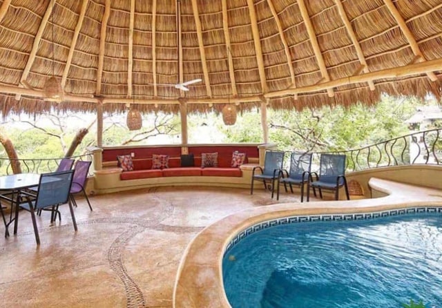 A spacious outdoor seating area features a thatched roof and comfortable seating along the perimeter. A swimming pool is visible, inviting relaxation. Nearby, a dining table accommodates several guests, surrounded by lush greenery that enhances the serene environment.