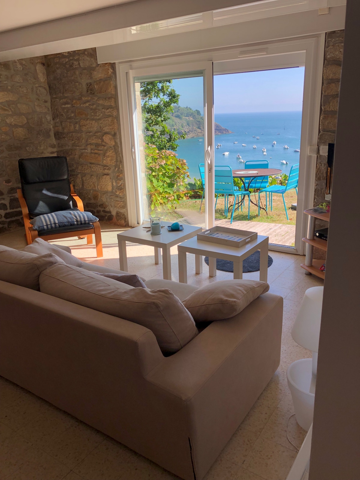 A seating area is presented with a light-colored sofa and modern table. Large glass doors offer a clear view of the sea and outdoor seating area with blue chairs. Natural light fills the space, highlighting the stone wall and serene ambiance.