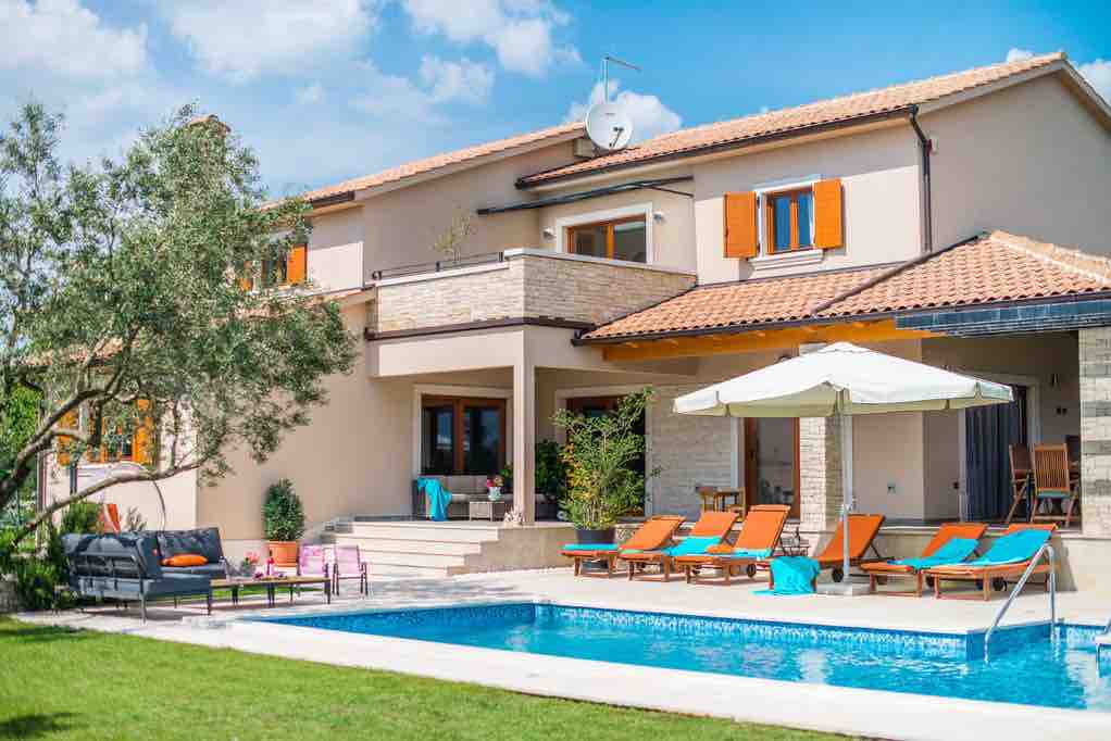 A modern villa with a large private swimming pool is showcased against a backdrop of blue skies. Sun loungers and an umbrella are situated beside the pool, while comfortable seating is arranged in a shaded area. Vibrant window shutters add a pop of color to the exterior.