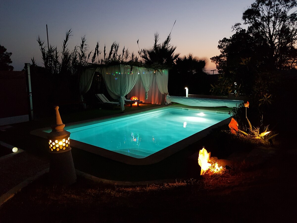 A serene pool area is illuminated at dusk, showcasing a clear, inviting water surface. Soft lighting from surrounding decorative elements enhances the tranquil atmosphere. A comfortable lounge chair rests nearby, framed by lush greenery that adds to the sense of privacy in the garden.
