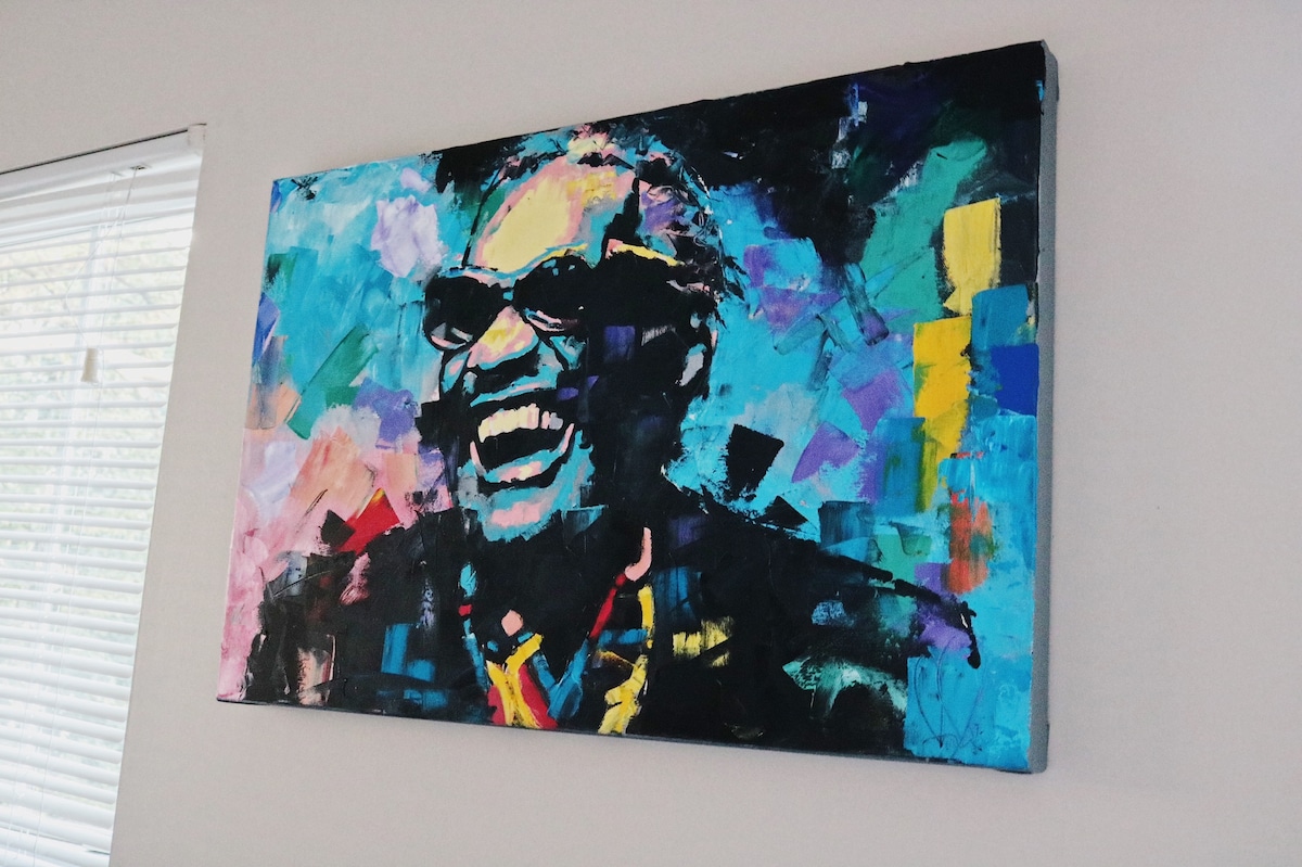 A vibrant painting of a smiling man adorns the wall, showcasing bold colors like blue, yellow, and pink. The figure is depicted wearing sunglasses, with a lively expression that adds character to the space. Natural light filters through nearby blinds, enhancing the artwork's visual impact.