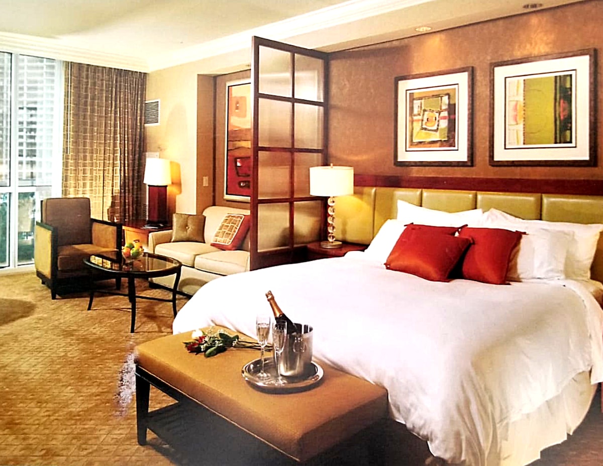 A spacious studio suite is shown with a king bed dressed in white linens. A seating area includes a pair of armchairs and a coffee table. Decorative wall art frames are visible, along with two bedside lamps casting a warm glow. Natural light filters through the large window.