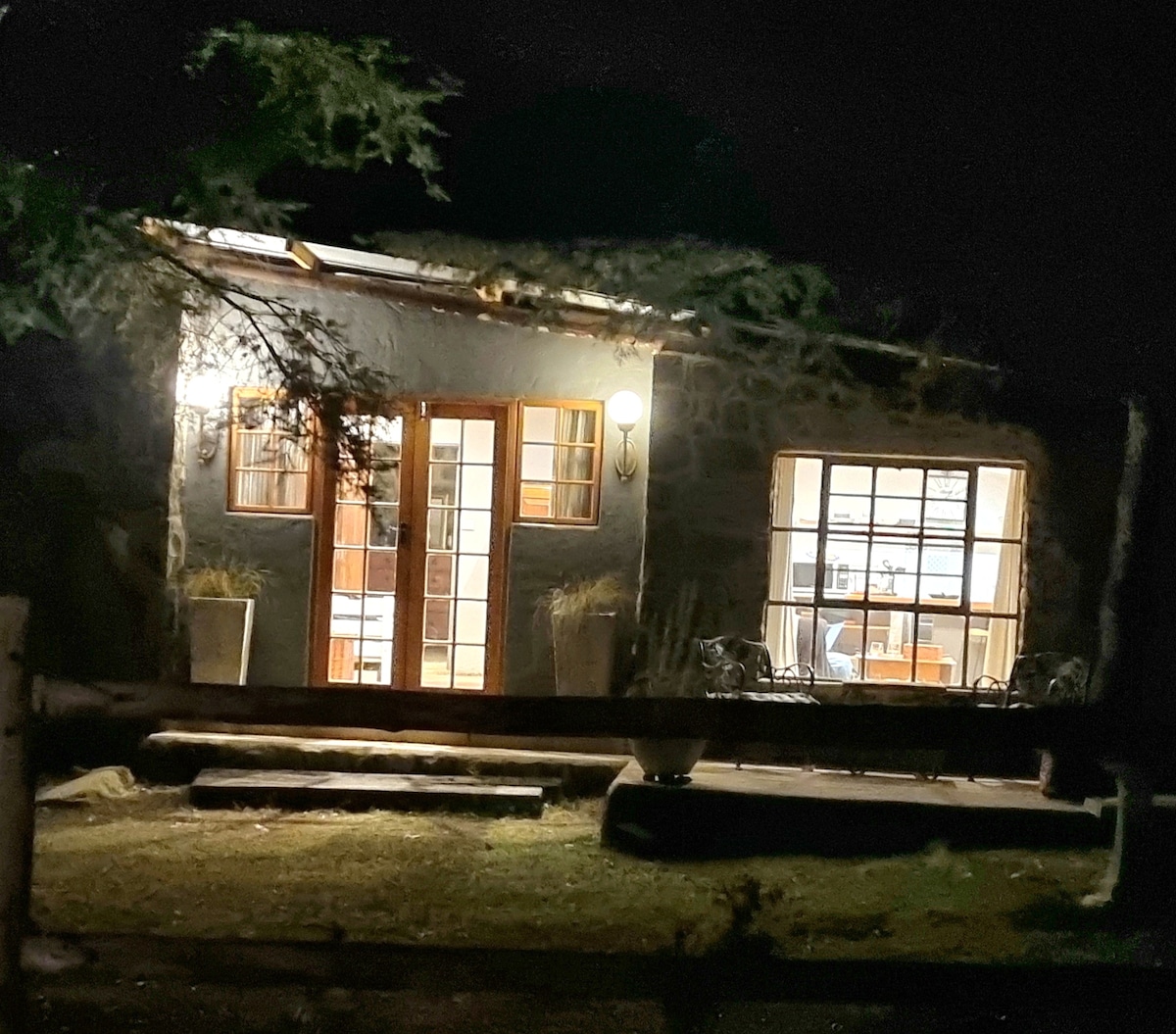 The exterior of a rustic stone barn is illuminated at night, revealing warm light spilling from the interior through large windows. A welcoming front door is flanked by potted plants, and native trees provide a natural frame to the peaceful scene.