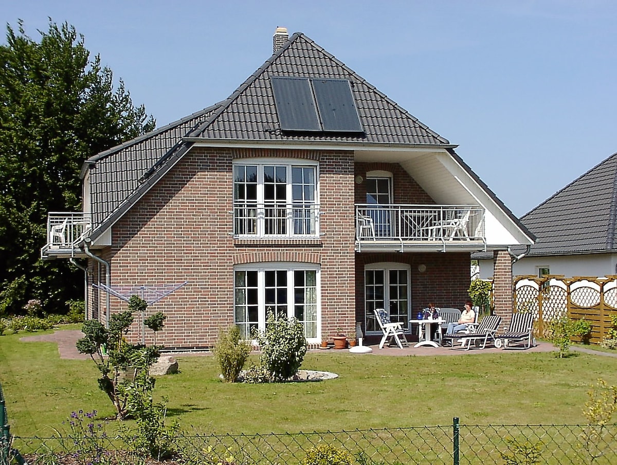 The charming two-story house is constructed with red brick and features numerous large windows. A balcony is visible on the upper level, while a patio set is arranged on the spacious lawn, surrounded by neat landscaping. Solar panels are mounted on the roof.
