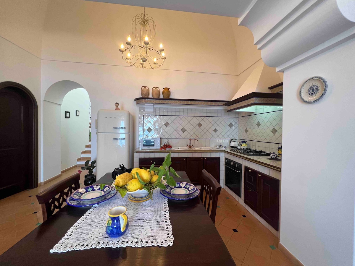 The charming kitchen features dark wood cabinetry and a decorative chandelier. A dining table is set with plates and a vase of lemons, while a vintage refrigerator and kitchen appliances are visible. The tiled floor adds to the inviting ambiance of the space.