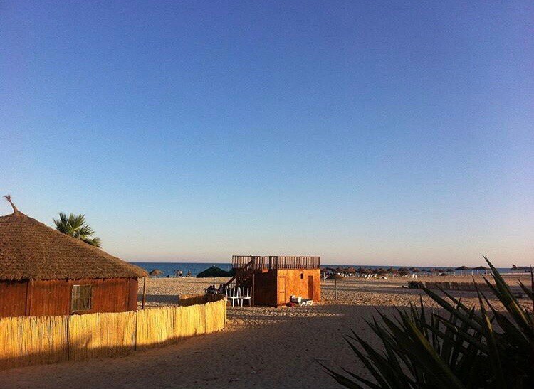 A beach scene is visible, featuring a wooden structure with a thatched roof, set against the backdrop of a clear blue sky. Soft sandy terrain leads to the shoreline, where people can be seen enjoying the beach in the distance.