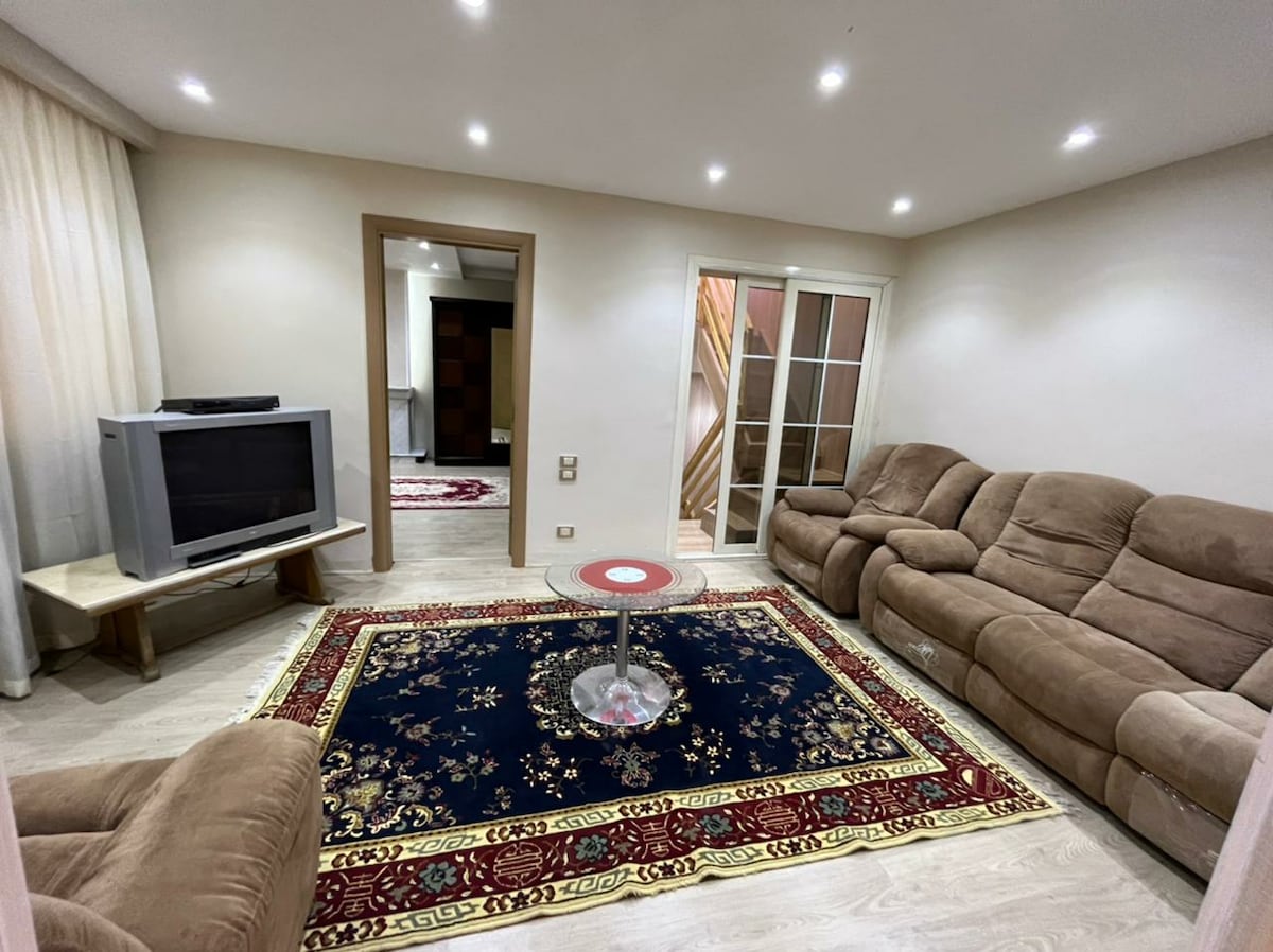 A living area features a plush beige sofa set arranged around a circular coffee table. A decorative rug with intricate patterns covers the floor. Soft lighting illuminates the room, highlighting the neutral-toned walls and providing a cozy ambiance.