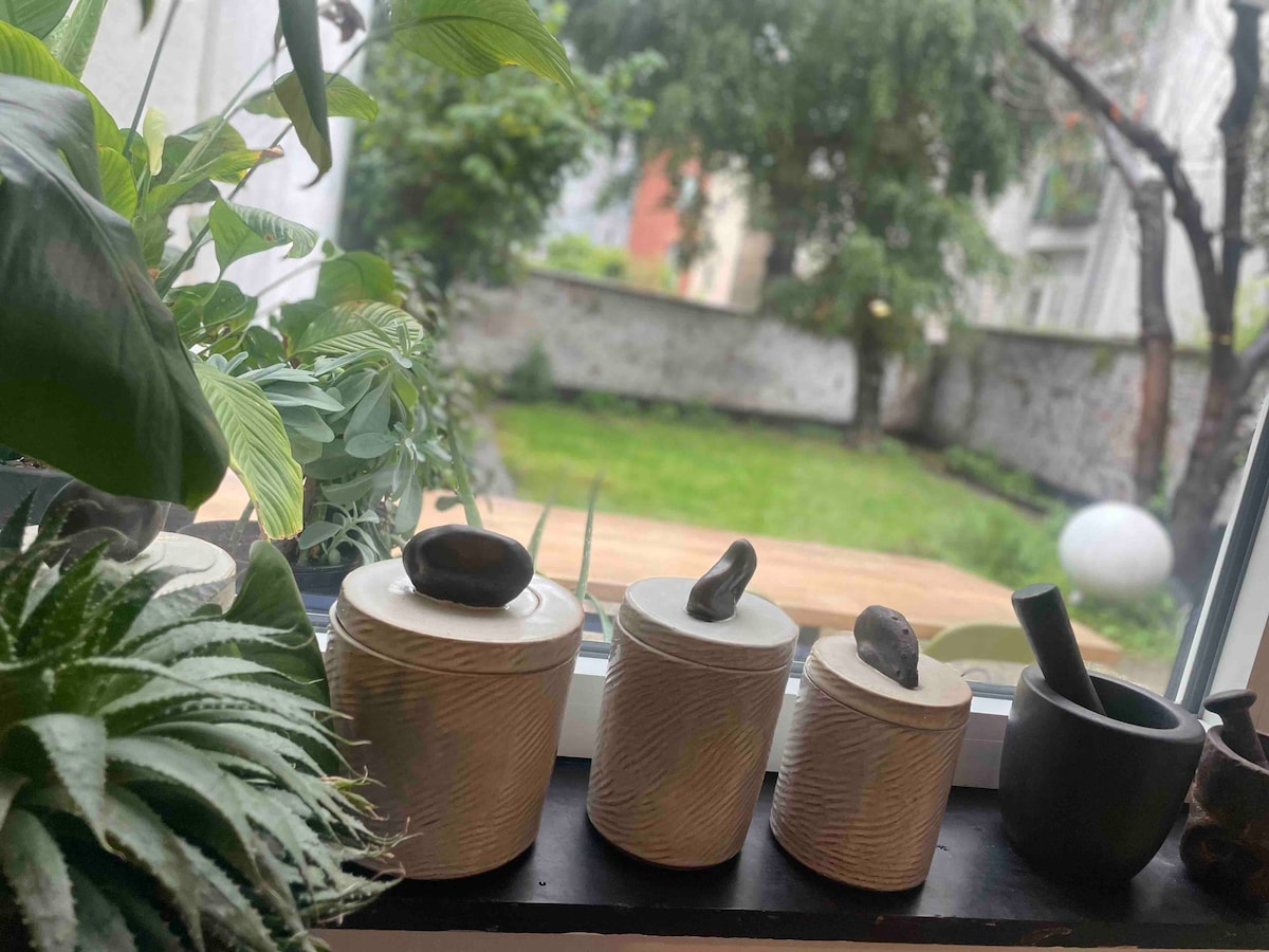 A view from inside the apartment showcases three decorative storage jars with natural stone lids, positioned on a windowsill. Lush greenery and potted plants frame the scene, leading to a vibrant garden visible through the window. A tranquil green lawn is complemented by wooden decking and nearby trees.