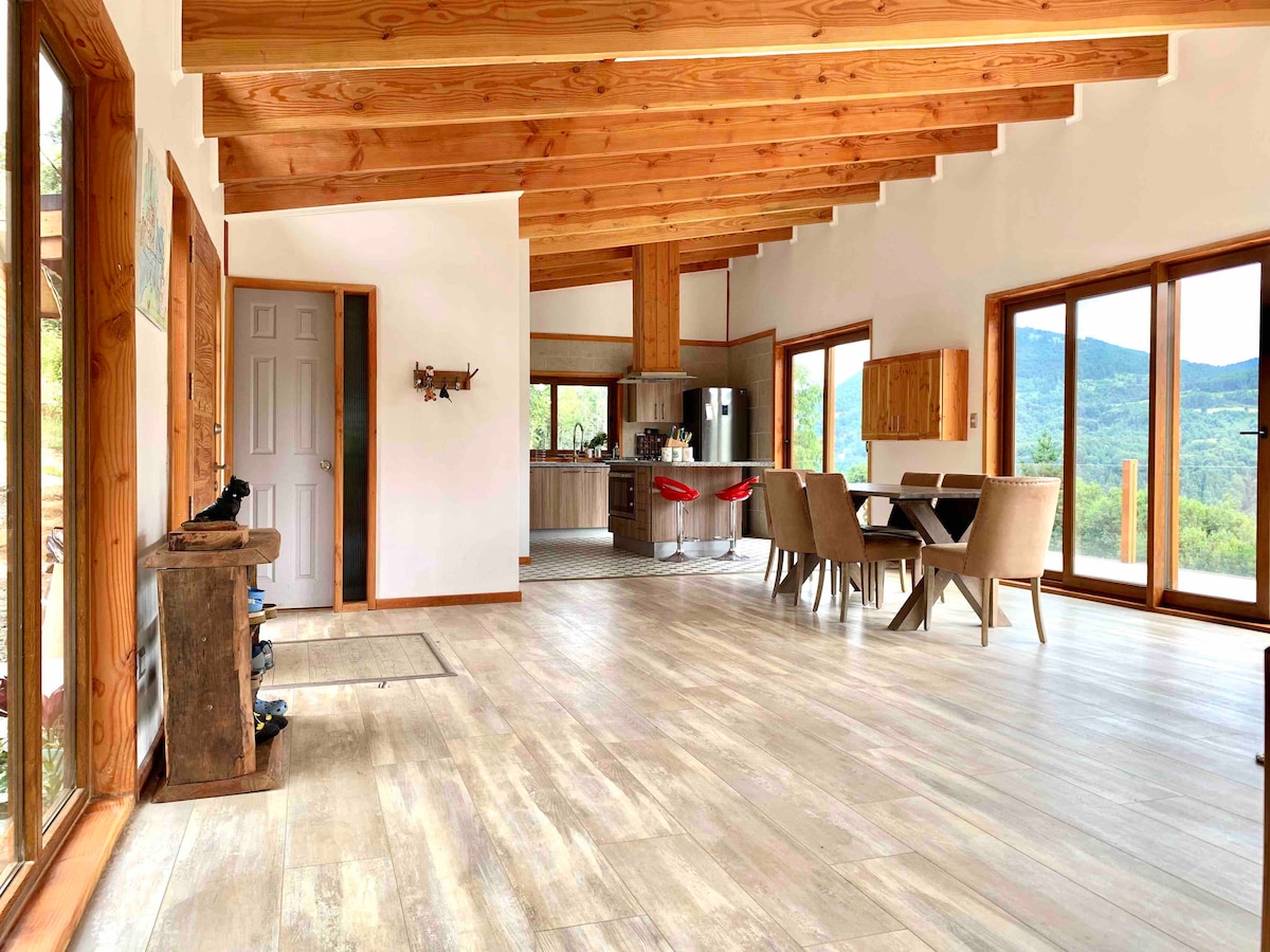 An expansive living area features large windows framing views of the native forested mountains. The space includes a dining table with chairs and a modern kitchen with an island. Wood beams stretch across the ceiling, enhancing the open and airy feeling.